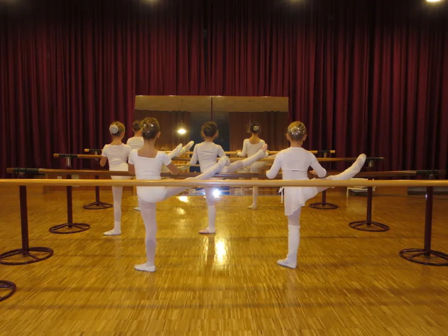 Ballettschülerinnen in weißen Trikots üben an der Ballettstange vor einem roten Vorhang